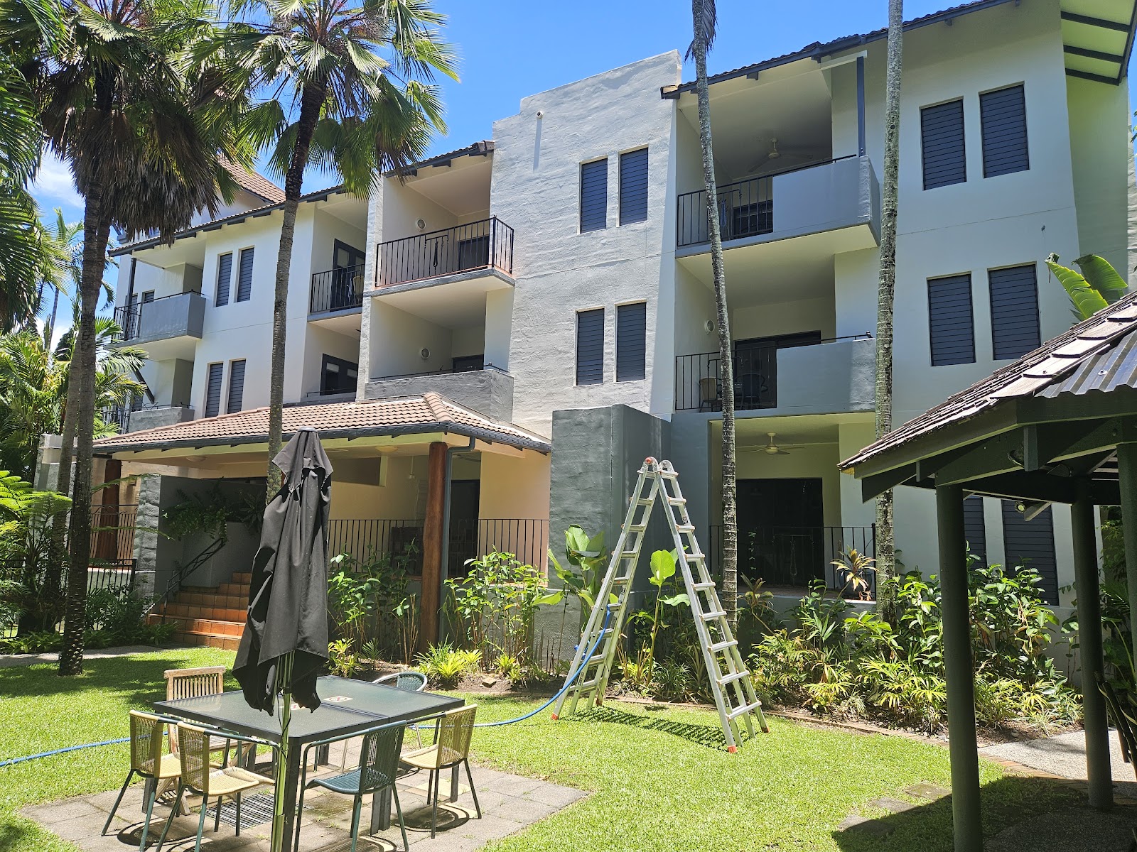 A three storey white apartment block with a ladder against the wall and a tropical garden in front